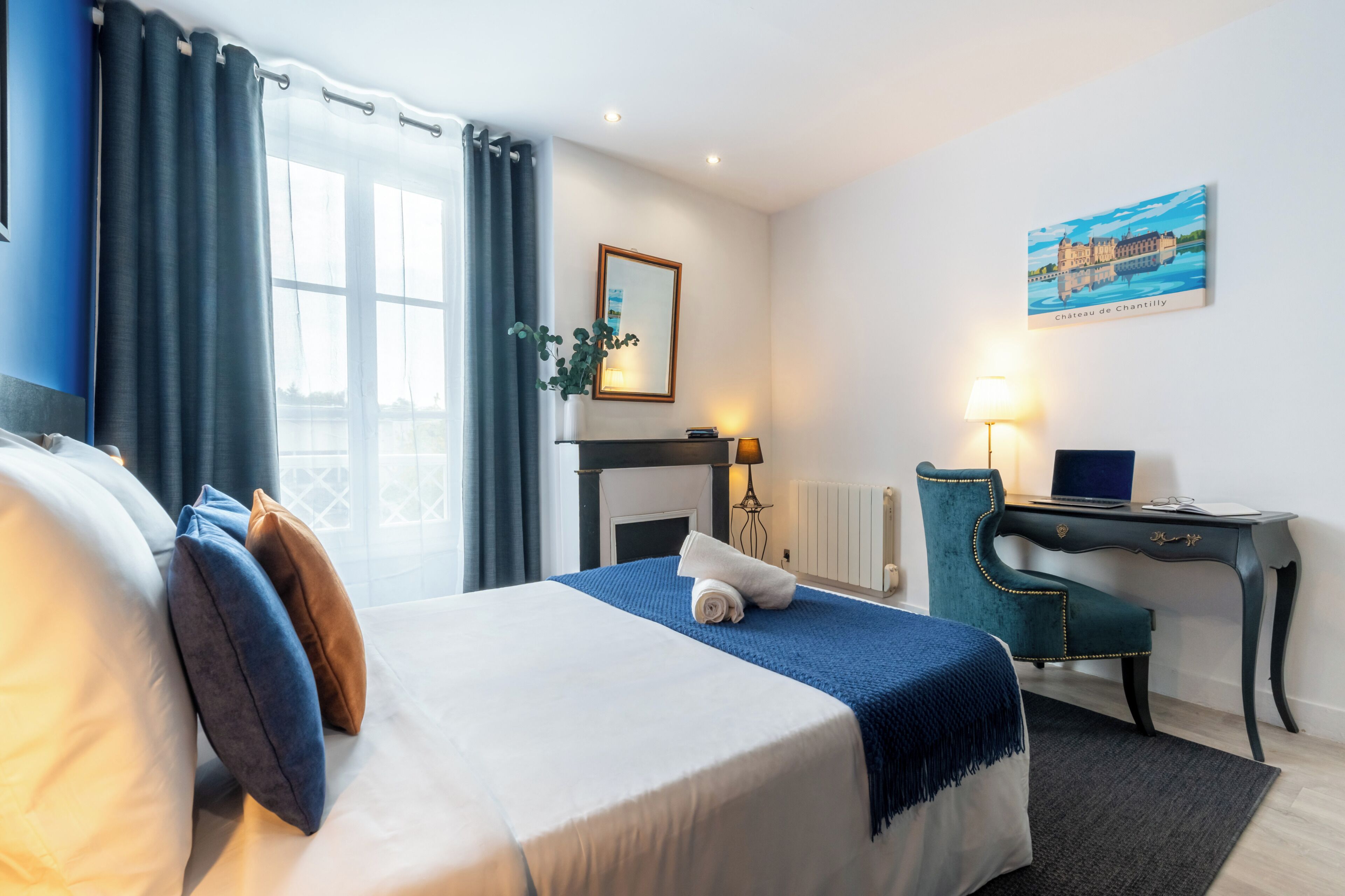A hotel room with a bed, a desk, a chair, a lamp, a painting, and a window with curtains.
