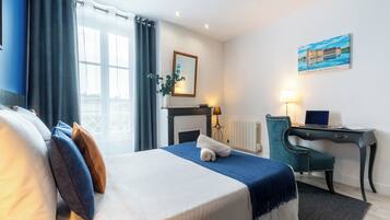 A hotel room with a bed, a desk, a chair, a lamp, a painting, and a window with curtains.