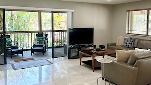 A spacious living room with a large TV, comfortable seating, and a balcony with a view of greenery.