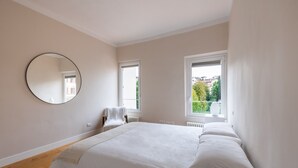 Une chambre avec un grand lit, un miroir rond, une chaise et deux fenêtres.