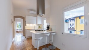 Une cuisine moderne avec des meubles blancs, un îlot central doté de tabourets et une grande fenêtre donnant sur les bâtiments environnants.
