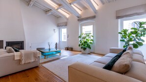 Un salon spacieux avec un plafond haut, des poutres en bois, une grande fenêtre, un canapé, une table basse et une plante en pot.