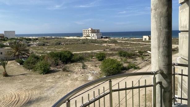 A view from a balcony overlooking a coastal area with a building in the distance, a sandy beach, and sparse vegetation. - Built in 2024, Sea view (sousse)