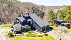 A large, modern two-story house with a covered patio, surrounded by a well-maintained lawn and a forested hillside.