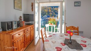 Een kamer met een houten kast, een televisie, een eettafel met een bloemen tafelkleed en uitzicht op een balkon met een tafel en stoelen.