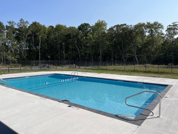Una piscina rectangular con escalera metálica y barandilla de seguridad, rodeada por una cerca negra y árboles.