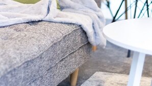 Un salon moderne avec un canapé gris, un coussin vert et une couverture blanche. On aperçoit en arrière-plan une table ronde blanche et une chaise noire.