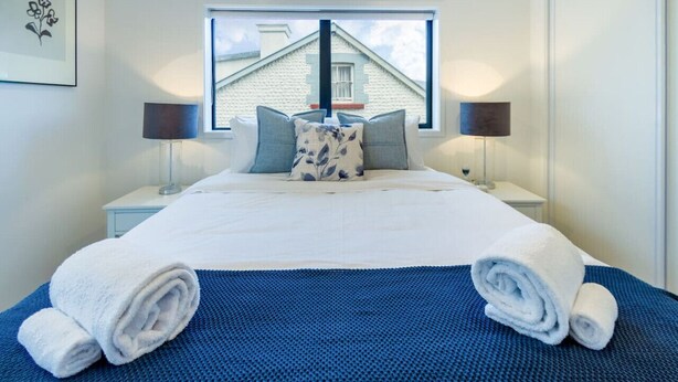 A neatly made bed with white linens and blue pillows, two bedside lamps, and a window with a view of a building. - Explore Addington and CBD 1 Bed w/Carpark (Christchurch)