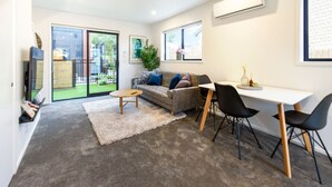 A modern living room with a grey carpet, a grey sofa with blue and brown cushions, a round wooden coffee table, a white desk with black chairs, and a large window with a view of a garden. - Explore Addington and CBD 1 Bed w/Carpark (Christchurch)