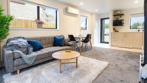 A modern living room with a grey sofa, a round coffee table, and a white shag rug. There is a dining area with a small table and chairs, and a kitchen area with wooden cabinets and a window. - Explore Addington and CBD 1 Bed w/Carpark (Christchurch)