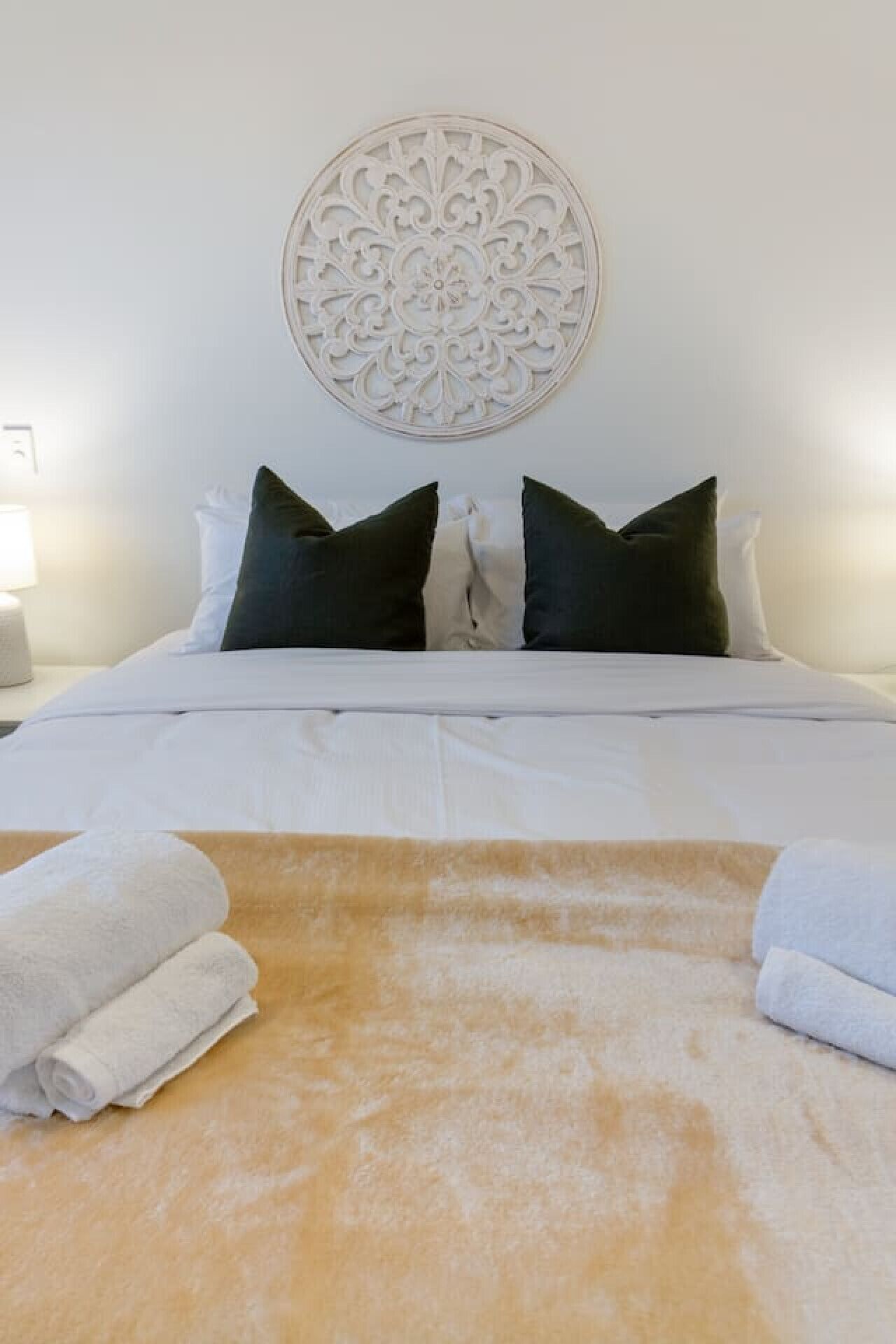 Een netjes opgemaakt bed met witte lakens, twee donkere kussens en een decoratief object aan de muur erboven.