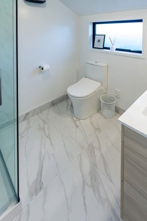 Une salle de bain comprenant des toilettes, un espace de douche vitré, un sol en marbre et une petite fenêtre ornée d’un cadre.