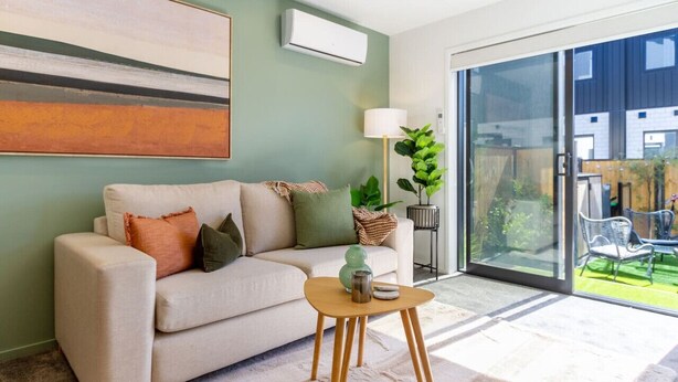 Een moderne woonkamer met een beige bank, een klein houten bijzettafeltje en een groot raam met uitzicht op een tuin en omliggende gebouwen.