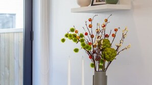 Een eetgedeelte met een witte tafel, twee stoelen, een vaas met takken en kaarsen, en een raam met uitzicht op een gebouw.