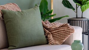Een woonkamer met een beige bank, een vloerlamp van hout, een groene plant en een tafeltje met een groene vaas en een metalen beker.