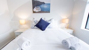 A neatly made bed with white linens and rolled towels, a blue pillow, and a painting of a coastal scene above it.