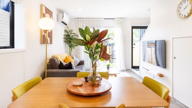 Un espace de repas moderne avec une table en bois, des chaises à coussins verts, un élément décoratif central et un canapé avec un plaid jaune en arrière-plan.