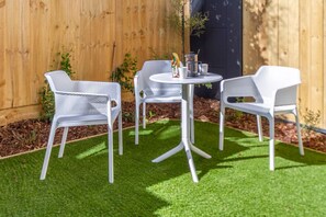 A small outdoor seating area with white mesh chairs and a round table, surrounded by artificial green grass and a wooden fence.