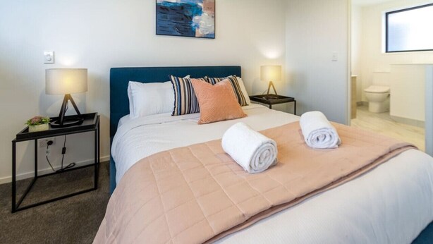 A hotel room with a bed, two bedside tables with lamps, a painting on the wall, and a bathroom visible in the background.