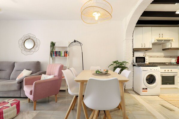 A modern living room with a sofa, armchair, dining table, and a washing machine in the background.