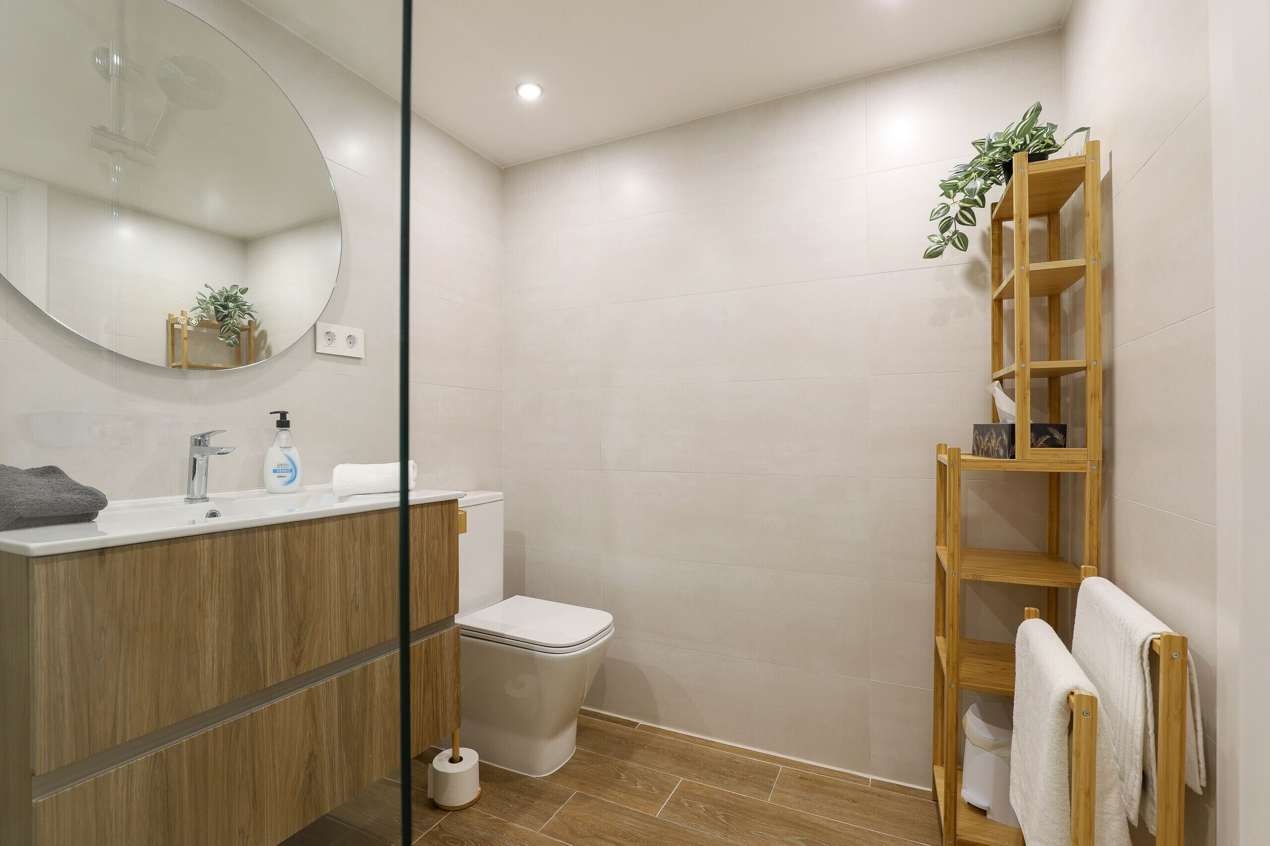 A modern bathroom with a wooden vanity, a toilet, a glass shower enclosure, and a wooden shelf with a plant.