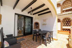 An outdoor patio with a table and chairs, a brick oven, and a view of the interior through a glass door.