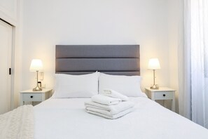 A neatly made bed with white linens and towels, two bedside lamps, and a grey headboard.