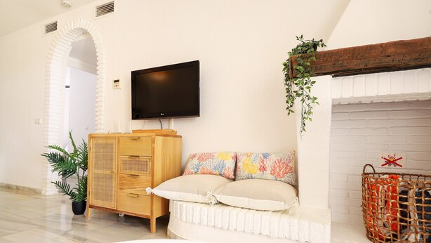 A living room with a flat-screen TV mounted on the wall, a wooden cabinet, a white sofa with colorful cushions, a white brick fireplace, and a wicker basket on the mantel. - Calahonda 2BED apartment - great location (Mijas Costa)