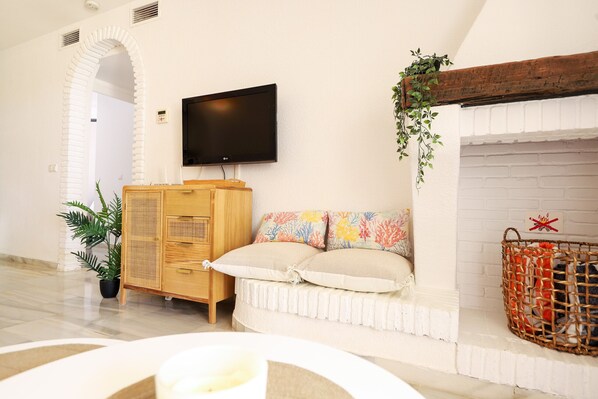 Ein Wohnzimmer mit flachbildfernseher an der Wand, einem Holzschrank, einem weißen Sofa mit bunten Kissen, einem Kamin aus weißem Mauerwerk und einem Korbkörbchen auf dem Sims.