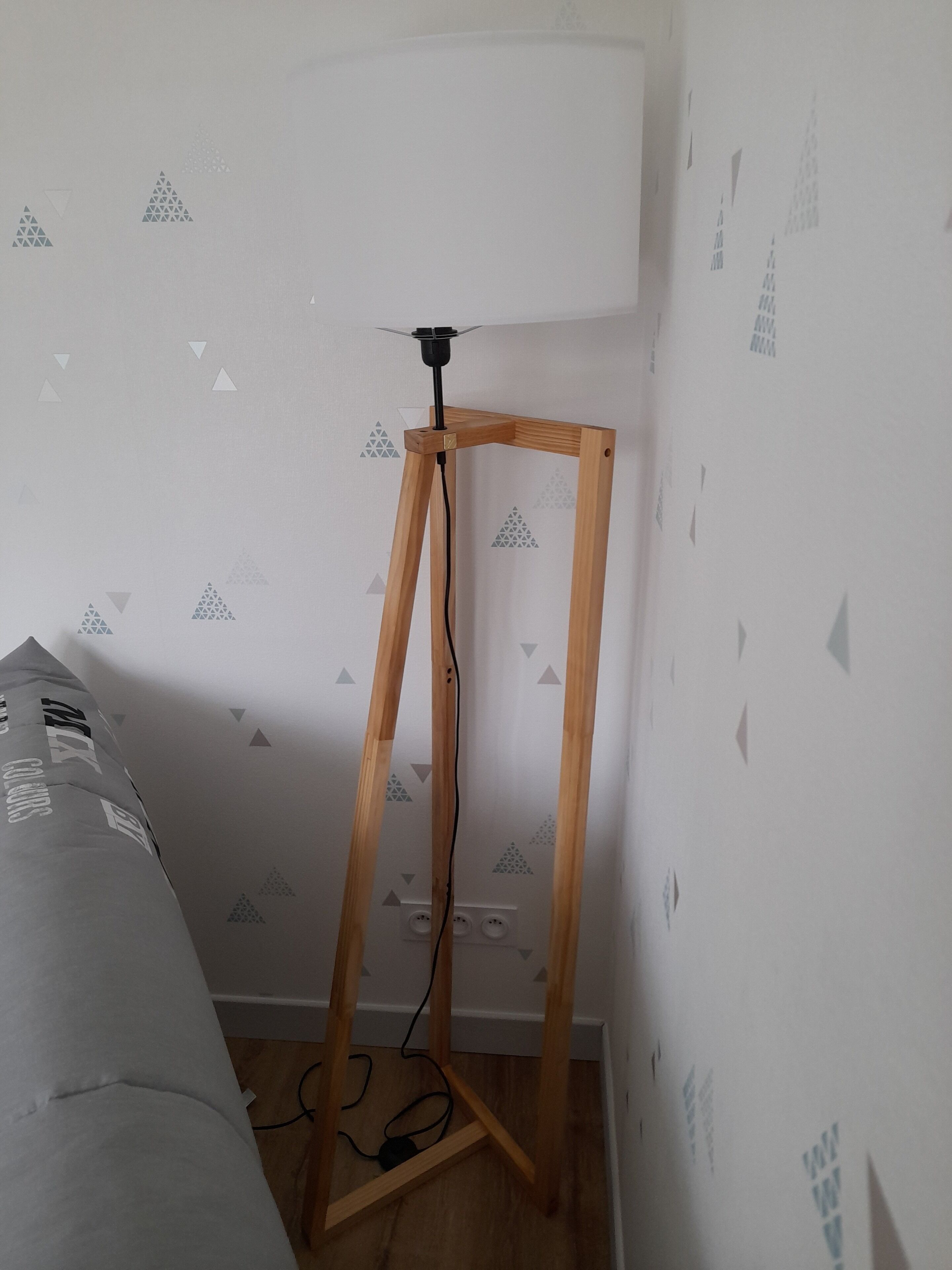 Habitación de esquina con una lámpara de pie sobre un atril de madera, una cama con cubrecama gris y una pared con diseño geométrico.