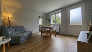 Een moderne woonkamer met een grijze bank, een houten eettafel en stoelen. Er is een lamp, een radiator en grote ramen die zorgen voor daglicht.