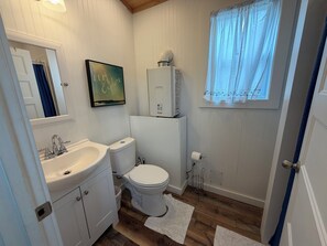 A bathroom with a white sink, toilet, and mirror, a framed picture on the wall, and a window with sheer curtains. - Tiny House on Center Hill Lake (Baxter)