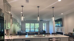 A modern kitchen with white cabinetry, a central island, pendant lighting, and a large window providing natural light. - Marvel Lake House Too (Hot Springs)