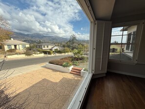 A view from a room with a large window overlooking a residential street, a landscaped garden, and distant mountains.