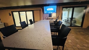Une salle de conférence avec une longue table et des chaises, un écran de télévision diffusant l’émission « The Game », et une vue sur un espace extérieur aménagé avec des tables et des chaises, accessible par des portes vitrées.