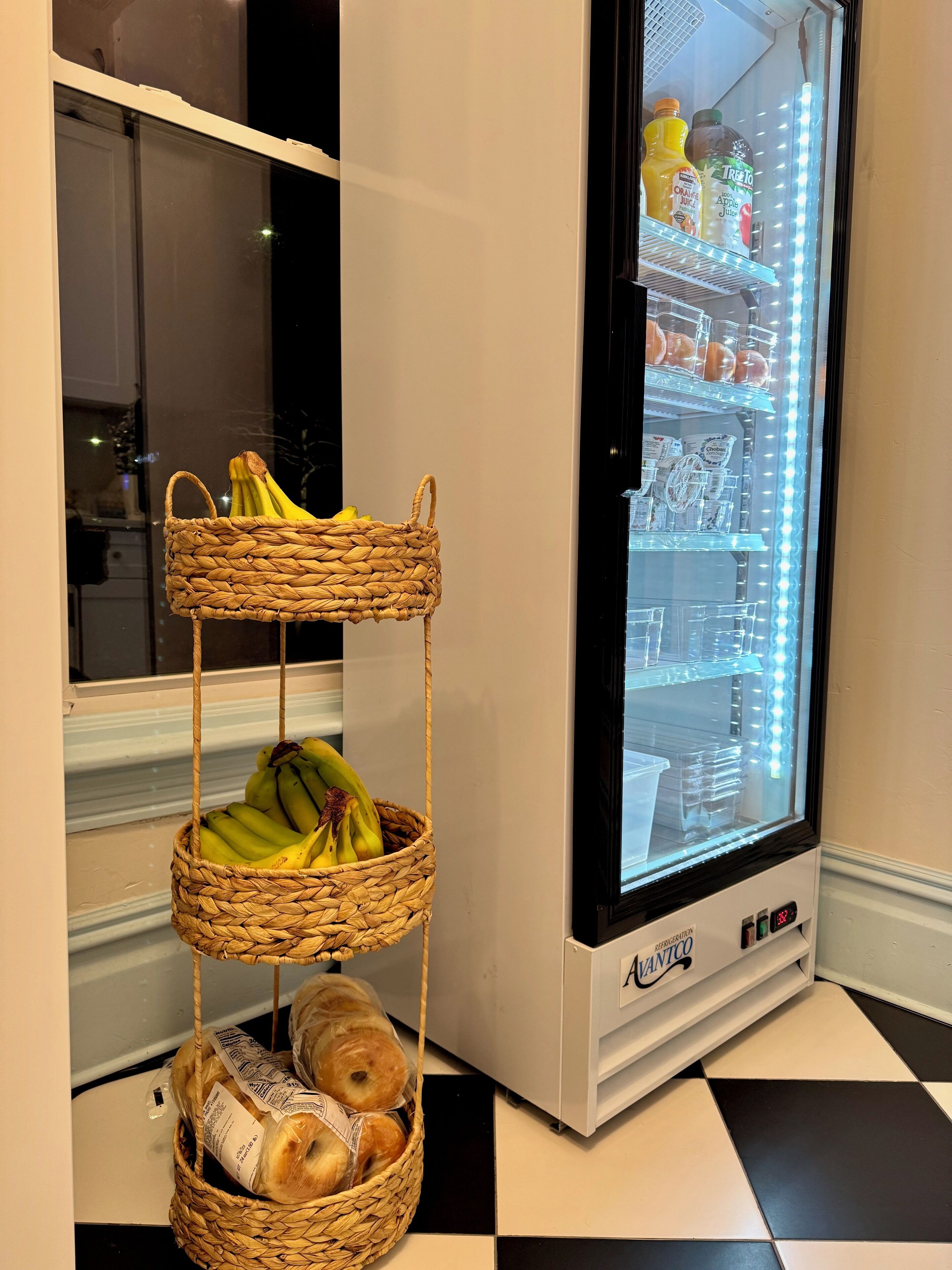 A refrigerator with a lighted interior, a wicker basket with bananas and bread, and a window with a view of the outside.