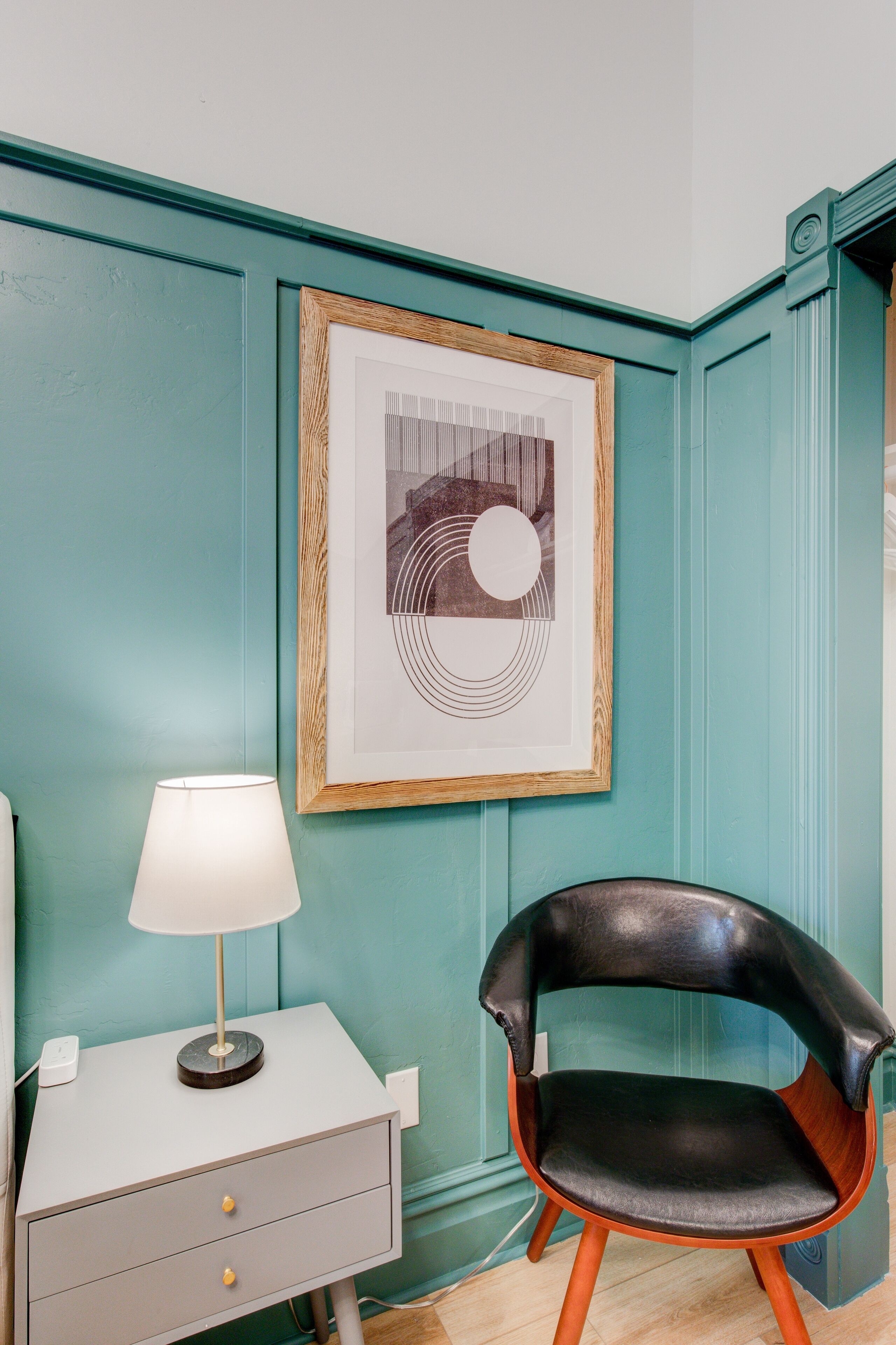 A teal-walled room with a framed abstract artwork, a white bedside table with a lamp, and a black leather chair.