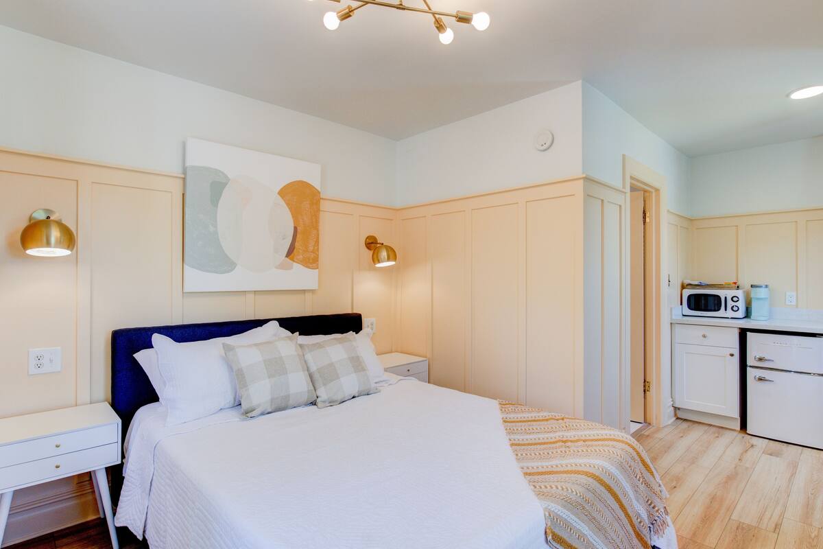 Camera da letto con un letto, comodini, un quadro astratto appeso al muro e un angolo cottura con forno a microonde.