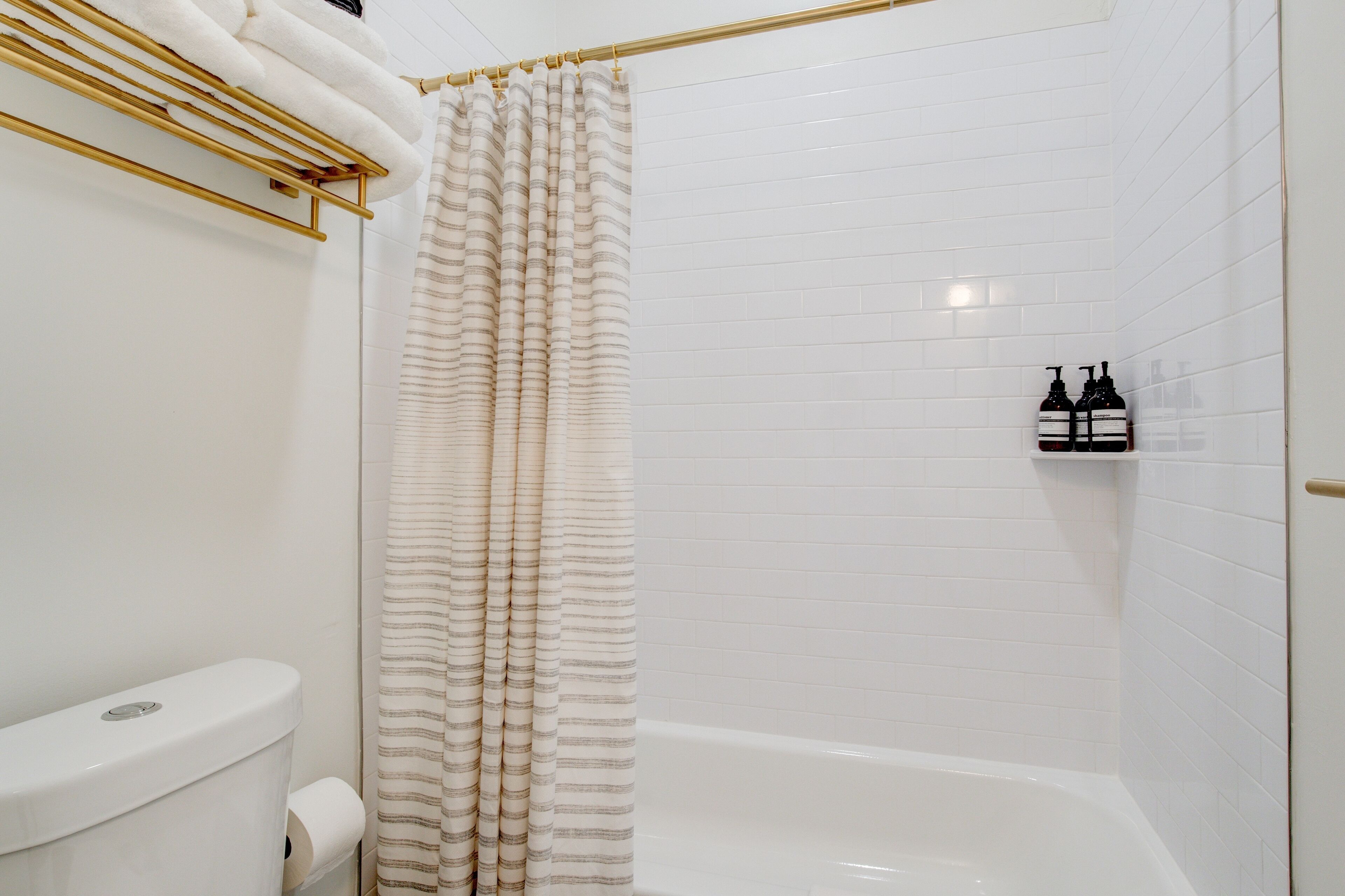 A bathroom with a white tiled wall, a white bathtub, a striped shower curtain, and two bottles of shampoo on a shelf.