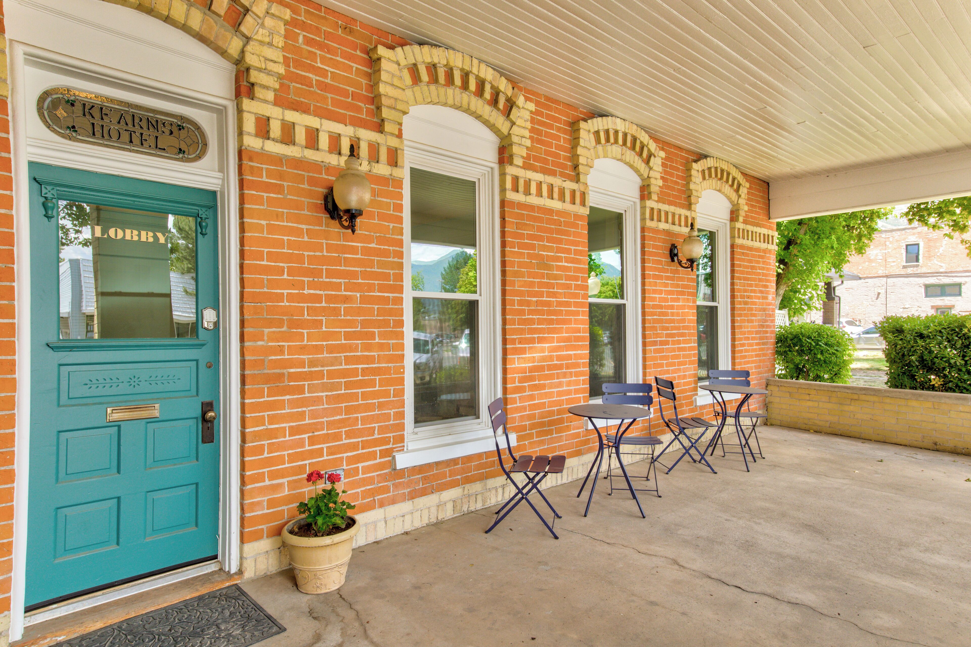 A brick building with a teal door labeled "Lobby" and a sign for "Kearns Hotel." There are outdoor chairs and a potted plant near the entrance.