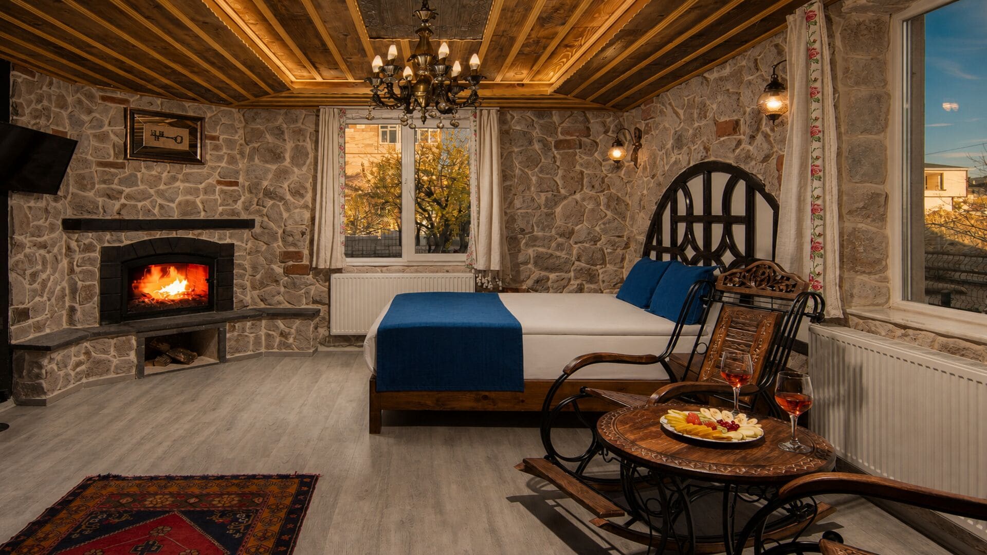 A cozy room with a stone fireplace, a bed with blue bedding, a dining table with fruit, and a view of trees through a window.