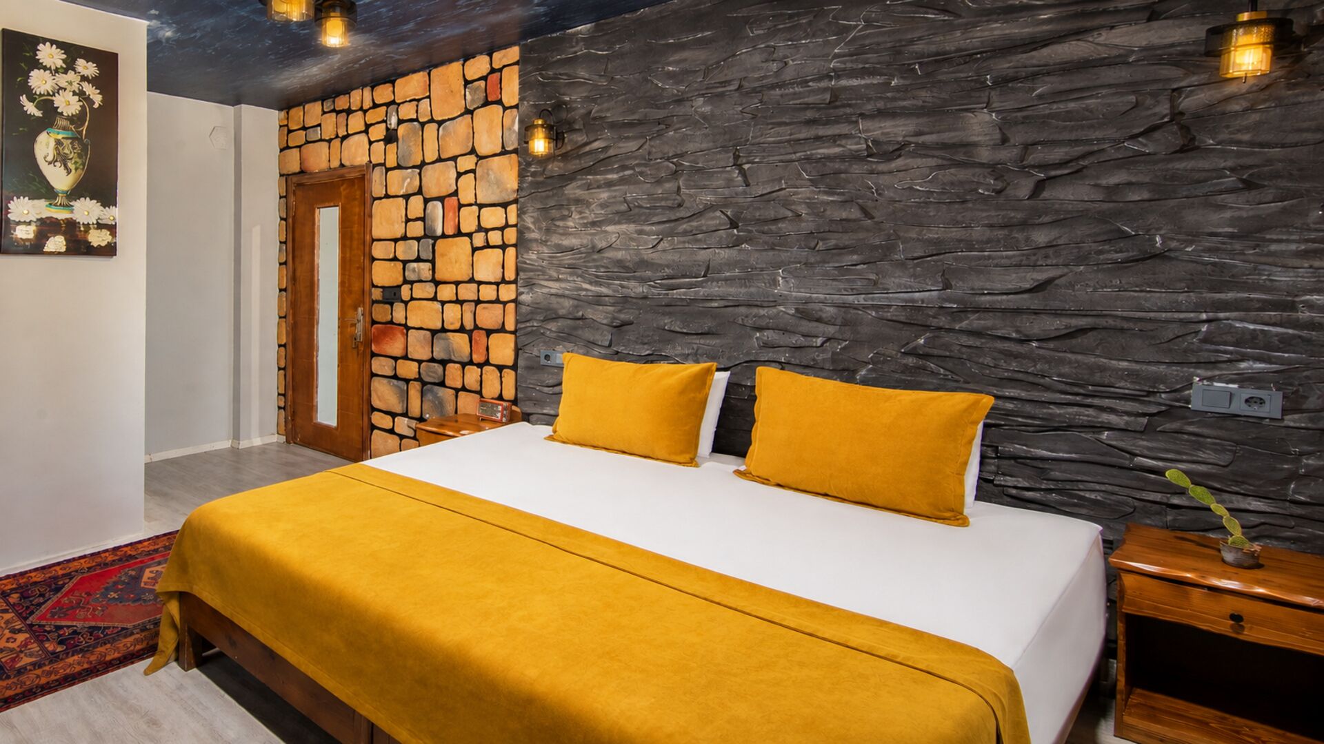 Une chambre avec un mur en pierre, un lit avec une literie jaune, une porte en bois et une table de chevet avec une lampe.