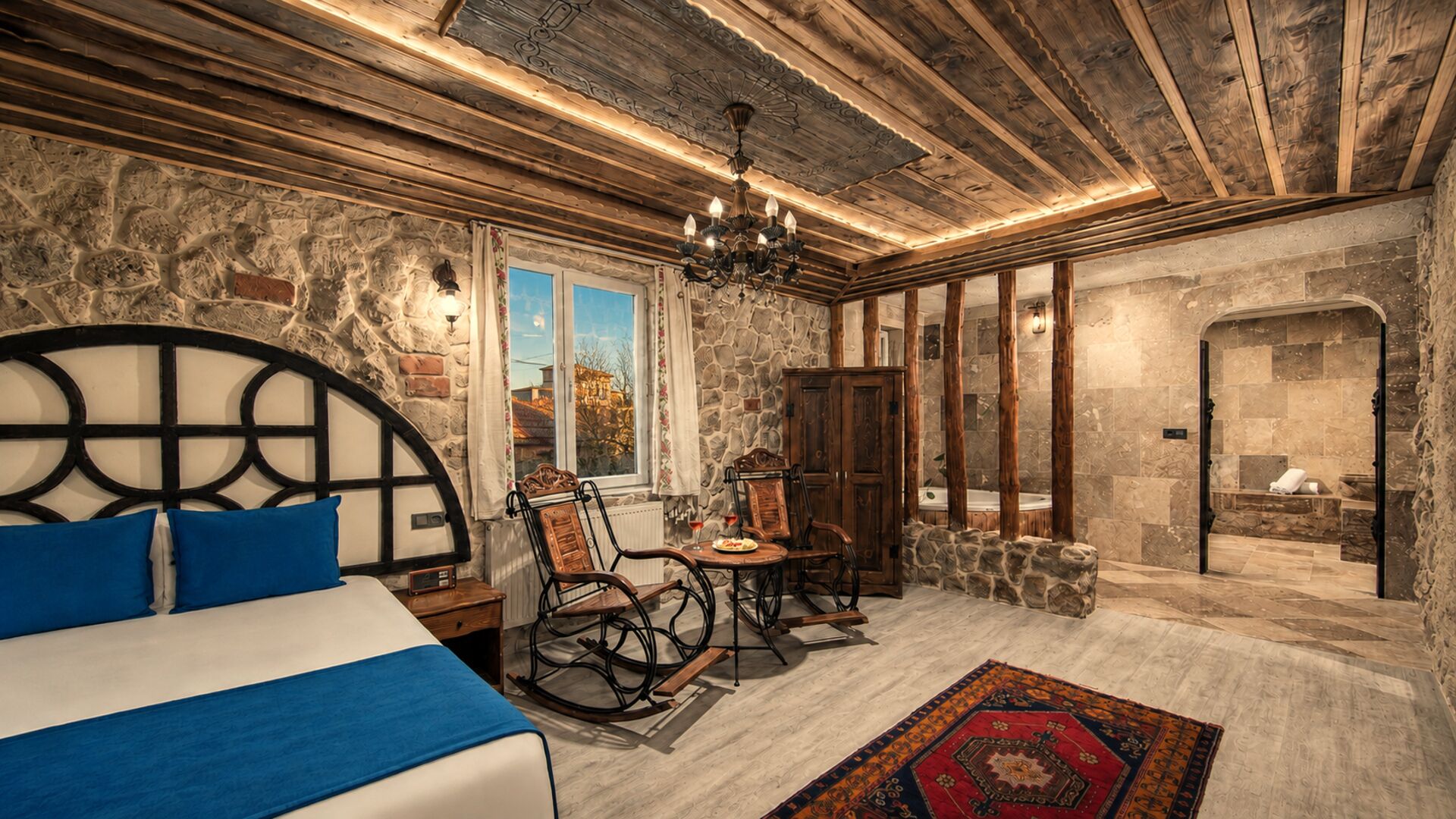 A room with a bed, a table, chairs, a chandelier, and a stone wall.