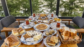 A table set with various dishes and drinks, likely for a meal, with a view of trees through large windows.