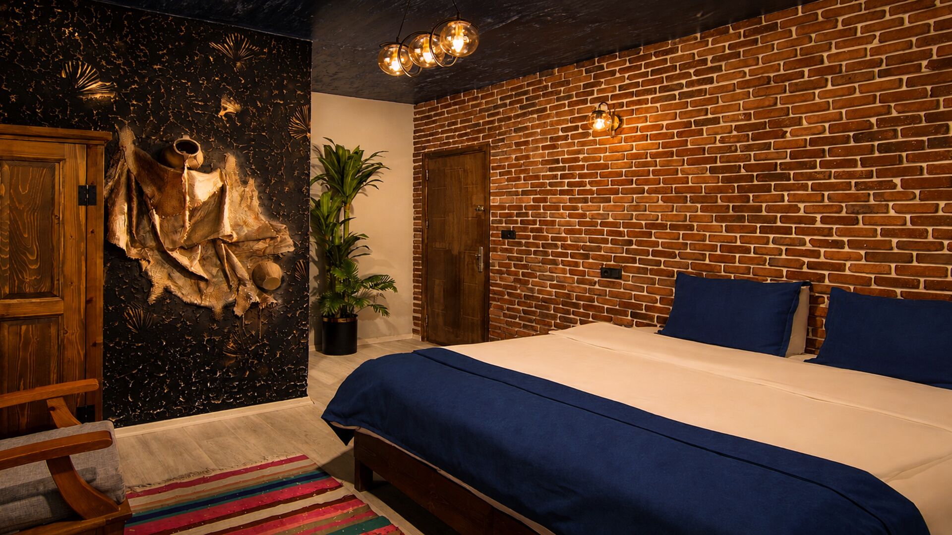A bedroom with a brick wall, a bed with blue bedding, a wooden door, and a plant.