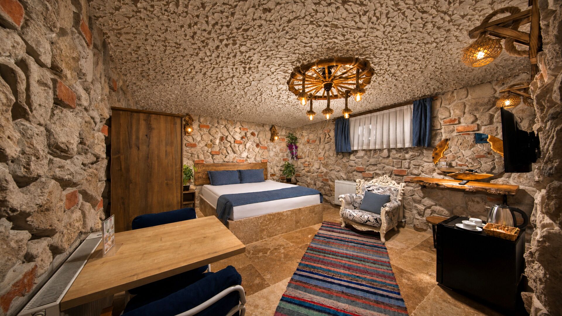 Una habitación rústica con pared de piedra, cama con ropa de cama azul, mesa de madera, alfombra estampada y una lámpara de techo original.