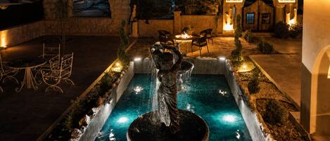 Un espacio exterior nocturno con una fuente central, muros de piedra y una zona de estar con mesas y sillas.