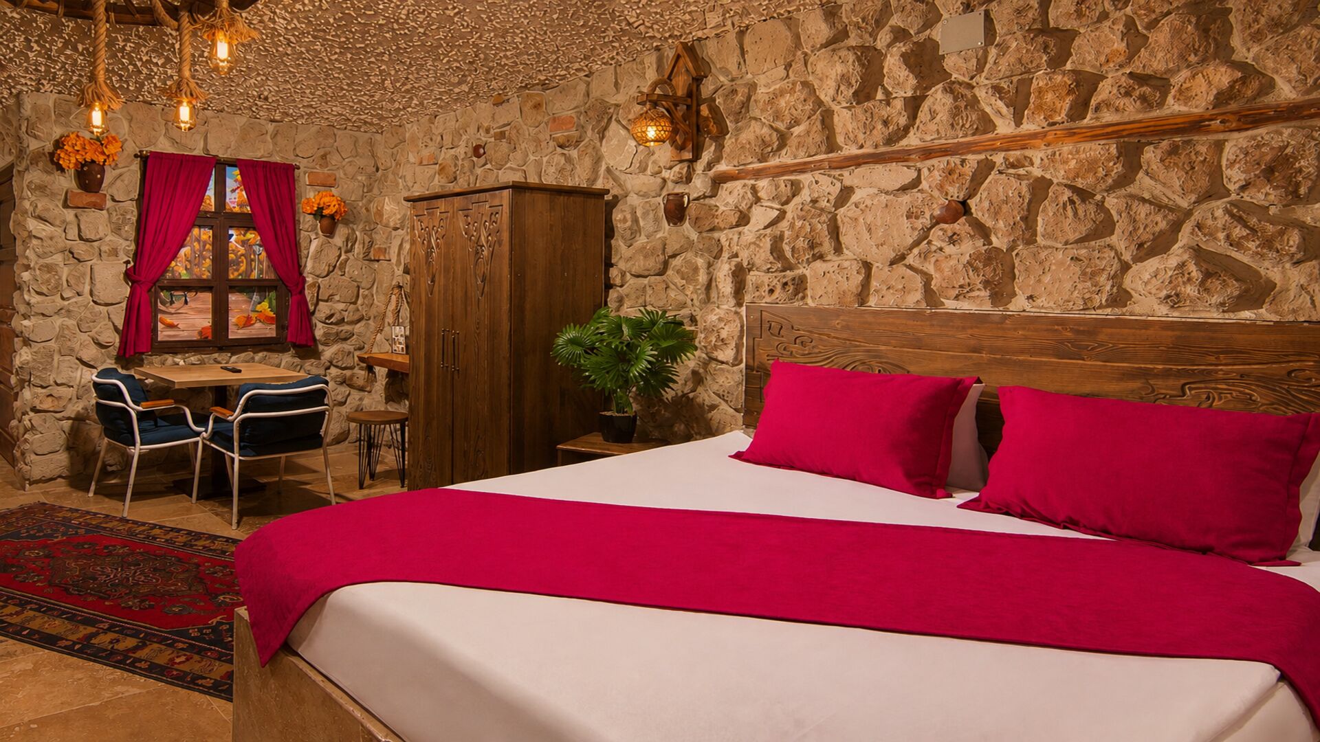 Un dormitorio con pared de piedra, cabecera de madera, cama con ropa de cama roja, escritorio con silla y lámpara colgante.