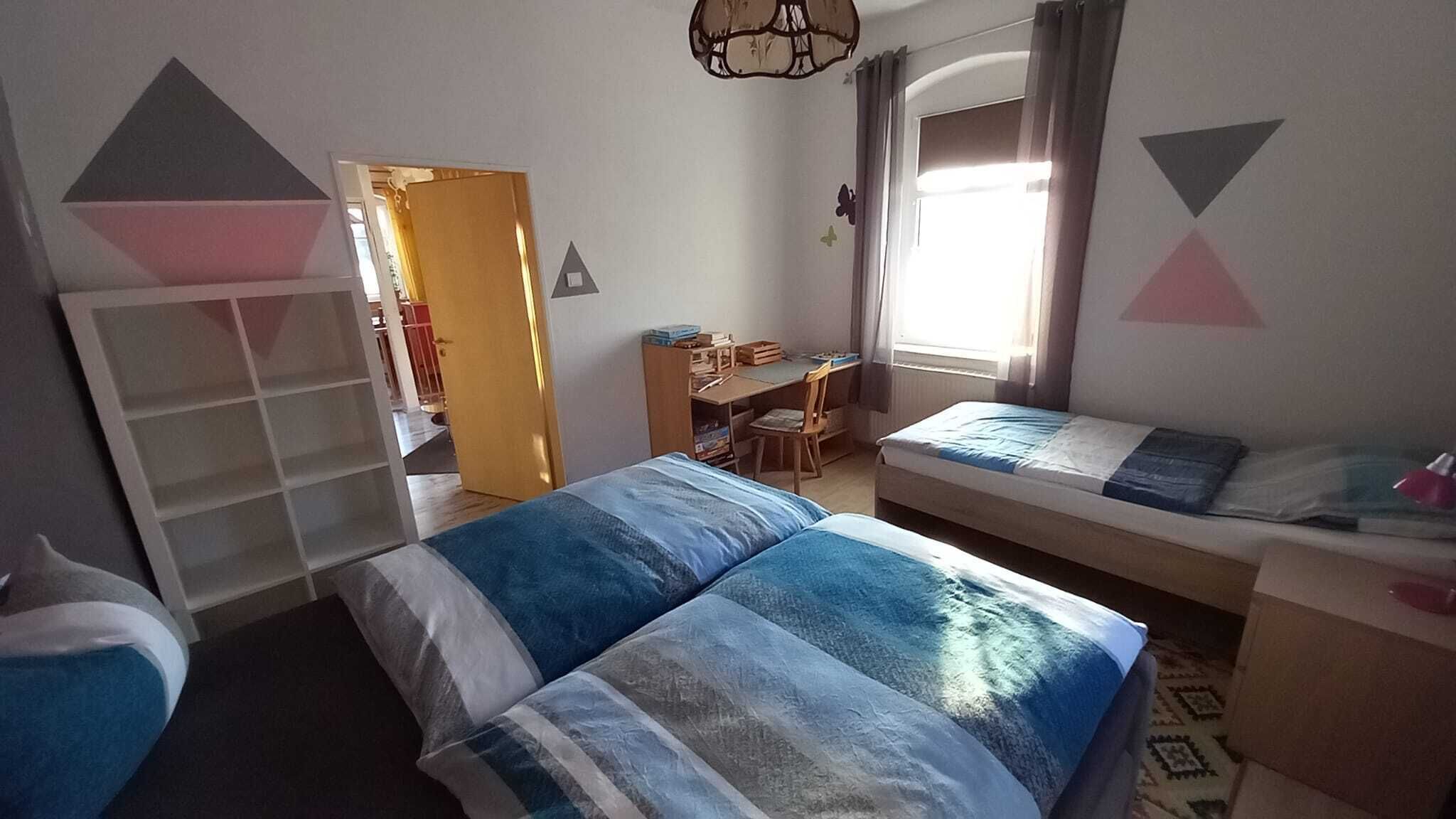 Ein Schlafzimmer mit Bett, Schreibtisch und Stuhl. Es gibt einen Kleiderschrank und ein Fenster mit Vorhängen.