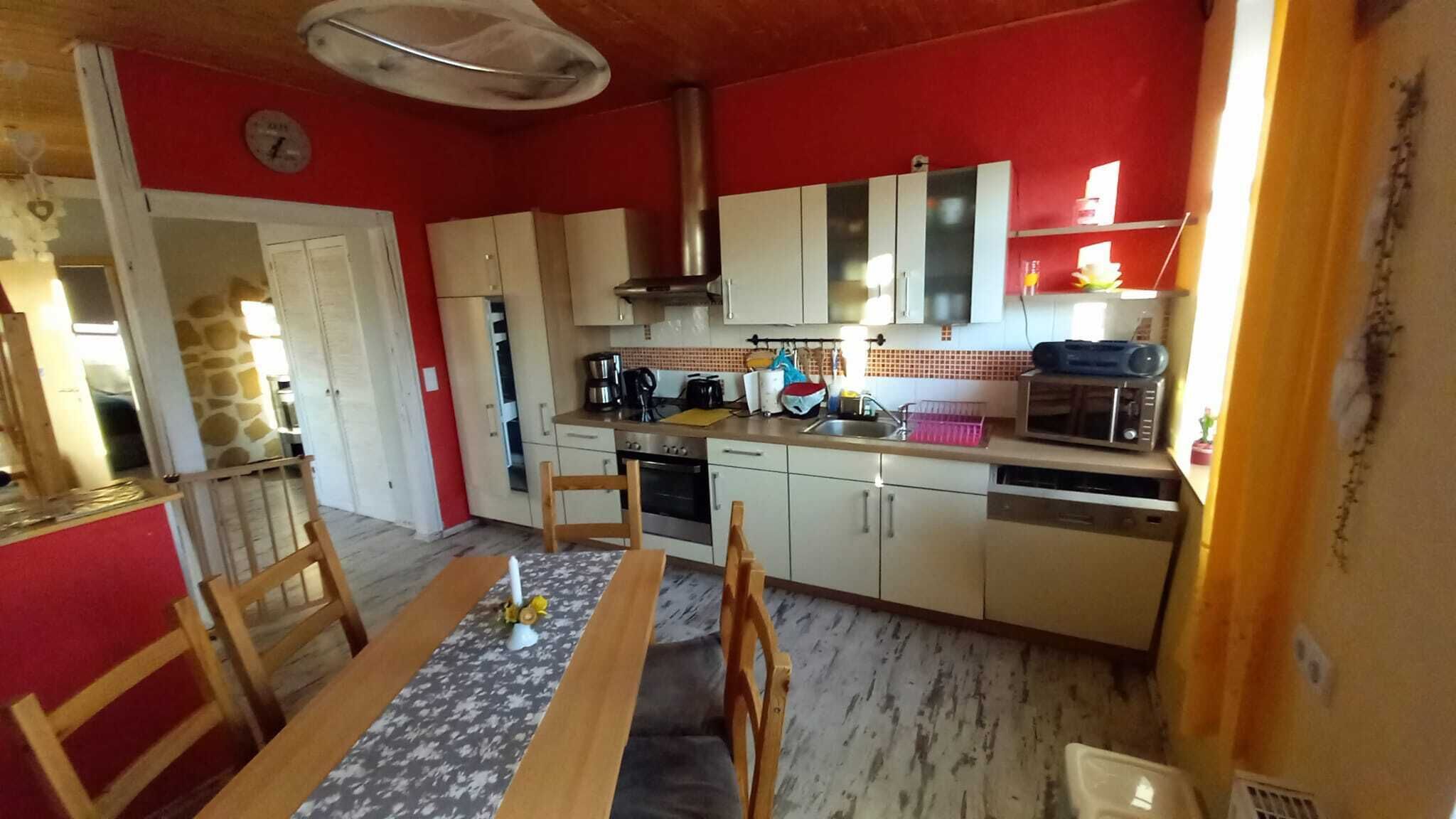 Een kleine keuken met witte kastjes, een magnetron, koffiezetapparaat en een spoelbak. Er staat een eettafel met stoelen en een klok hangt aan de muur.